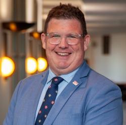 A headshot of Glen Morrison smiling at the camera, wearing a blue suit and blue tie.