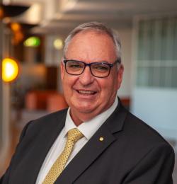 A headshot of Peter Maher smiling at the camera wearing a black suit and yellow tie.