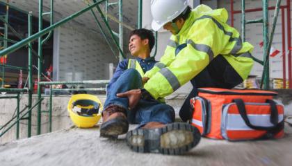 Two people on a work site, one is lying down in pain, the other is administering first aid