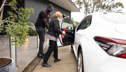 An image of Alith Jok assisting a SJQ client into a vehicle, he is wearing full uniform and smiling at the client.