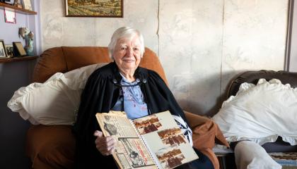 An image of Ann Demaine sittign in her home, holding an old photo album containing her professional recognitions.