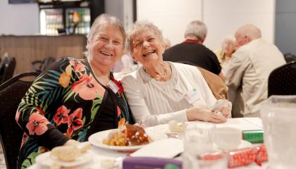 An image of two elderly women smiling happily at the camera while sat at a Christmas dinner table.
