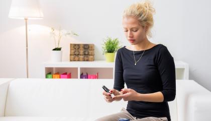 An image of a woman sat on a sofa, doing a diabetes blood sugar test on her index finger.