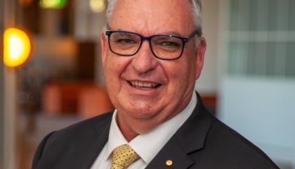 A headshot of Peter Maher smiling at the camera wearing a black suit and yellow tie.