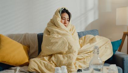 An image of a woman wrapped in a blanket on the sofa, surrounded by flu medicine.
