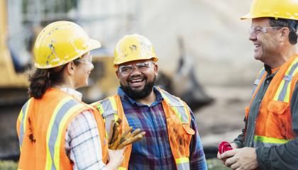 A group of three multi-ethnic workers at a construction site wearing hard hats, safety glasses and reflective clothing, smiling and conversing.