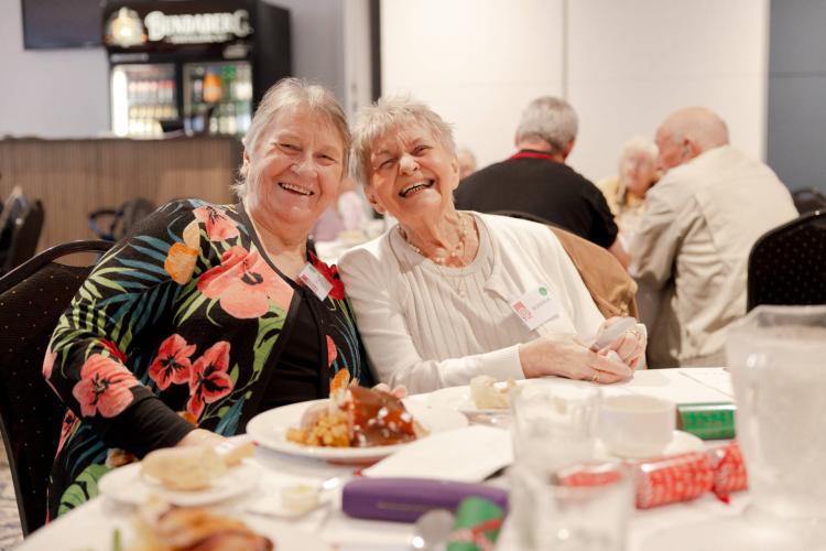 An image of two elderly women smiling happily at the camera while sat at a Christmas dinner table.
